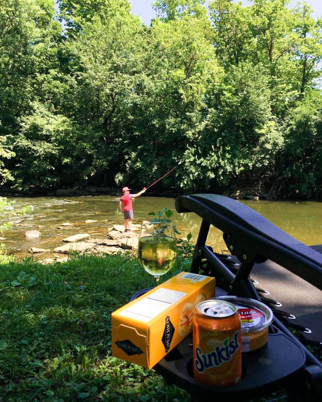 A picnic table featuring a can of soda and a box of chips, with a man fishing in the background.